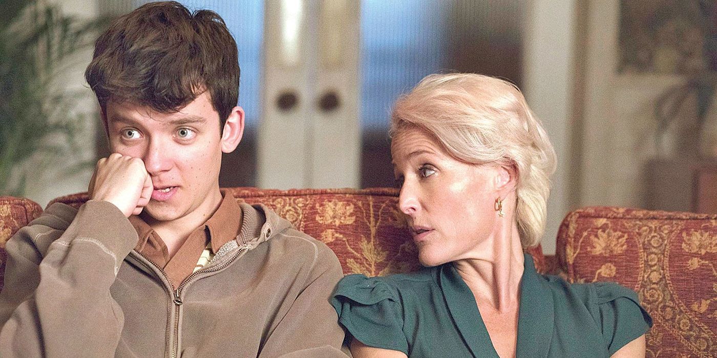 Otis (Asa Butterfield) looks on while holding hands and cheek to cheek with Jean (Gillian Anderson) while sitting on the couch in Sex Education.