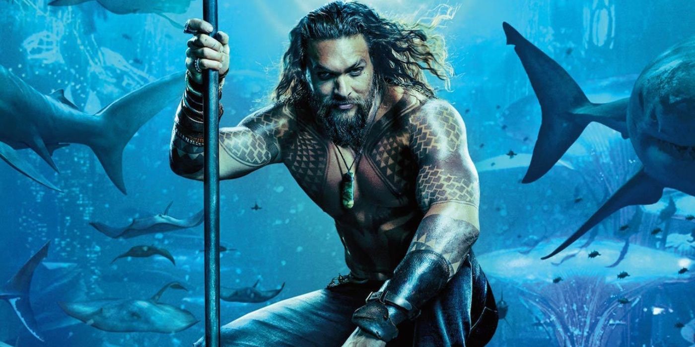 Jason Momoa as Aquaman kneeling underwater while holding a staff with sharks in the background in Aquaman