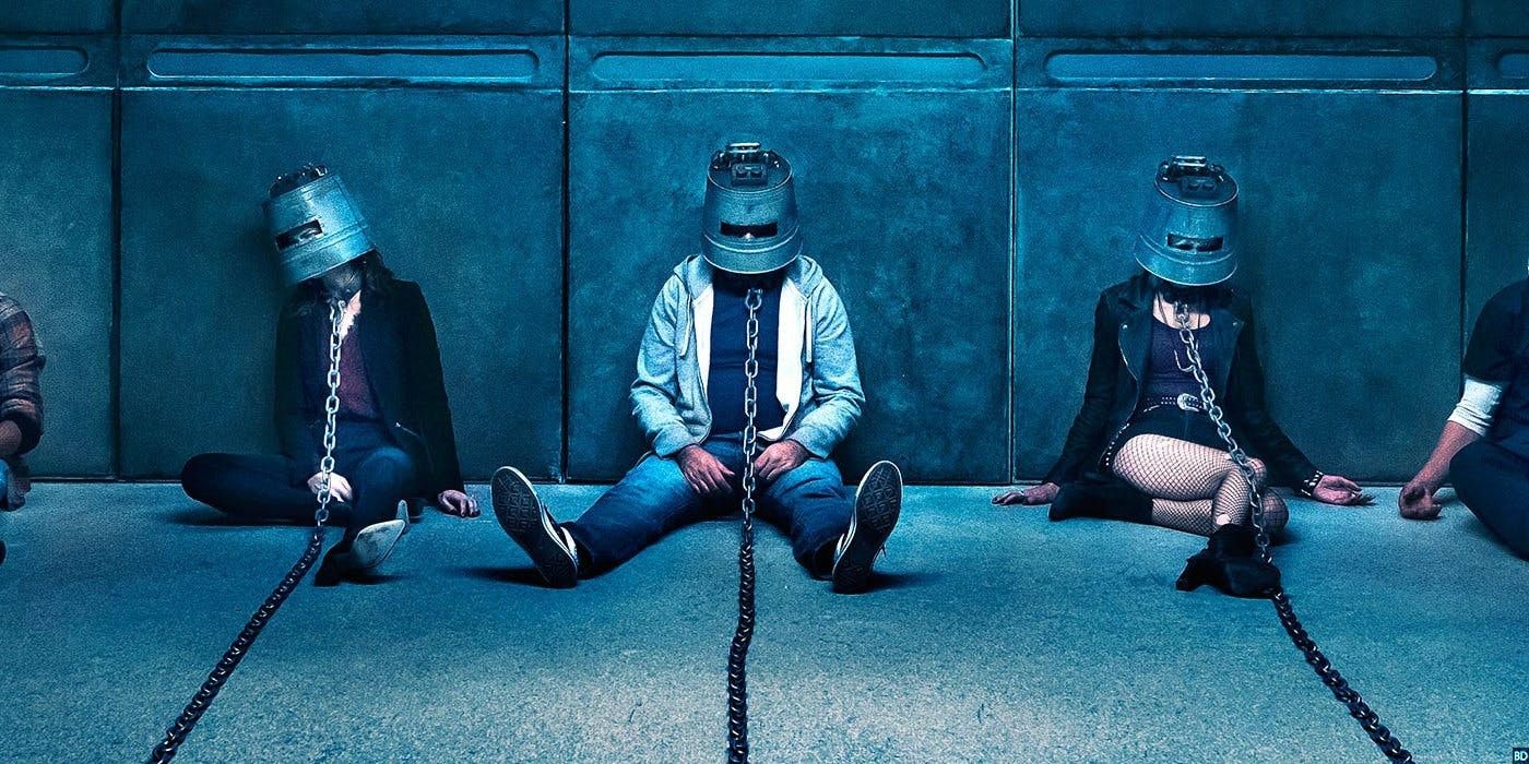 People sit on the floor wearing metal helmets chained to something offscreen in 'Jigsaw'.