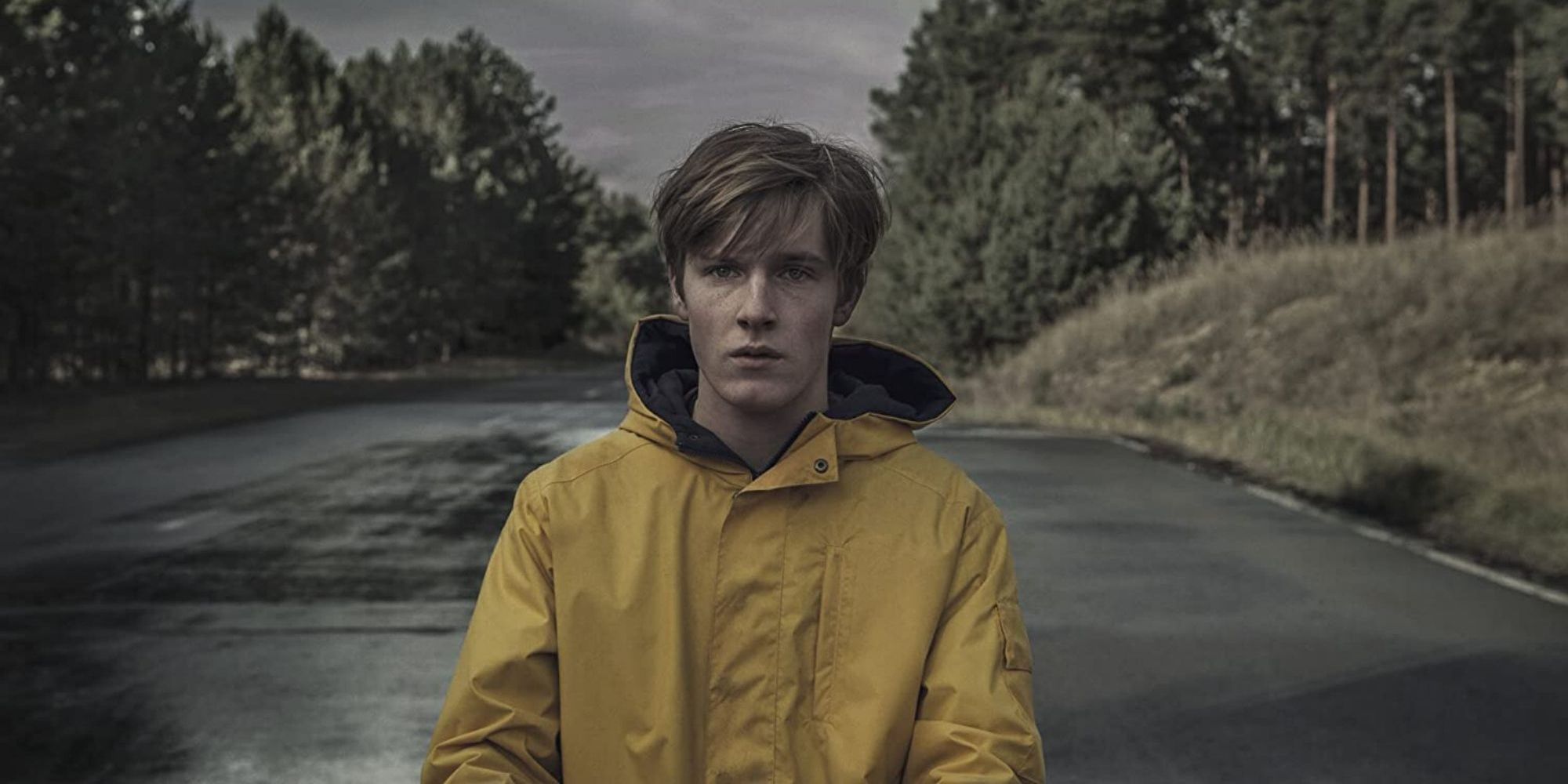 A young man in a raincoat stands on a road in Dark