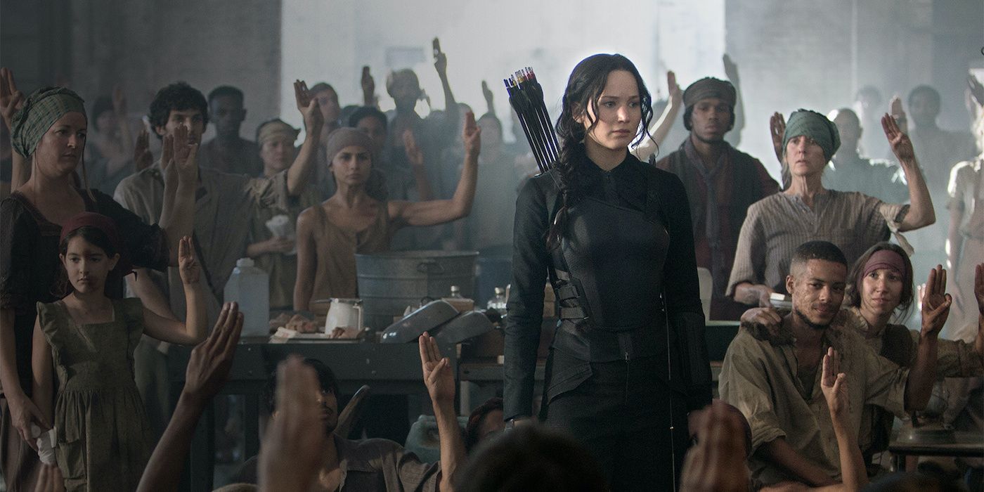 Katniss (Jennifer Lawrence) standing amongst a crowd of Panem residents who are holding up the three finger salute of resistance in The Hunger Games: Mockingjay Part 1