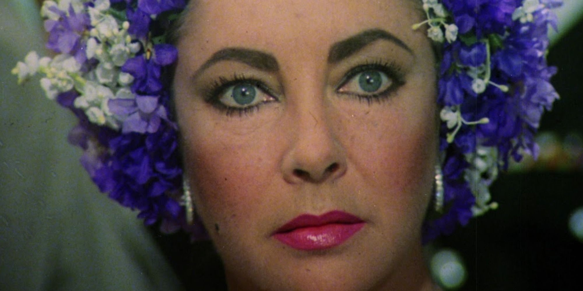 Marina Gregg (Elizabeth Taylor) wears a purple flowers in her hair. She is looking at someone with rage in her eyes.