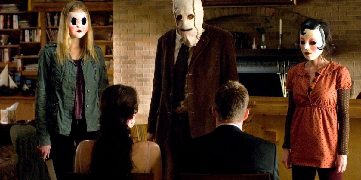 3 masked killers from The Strangers stand in front of a seated couple