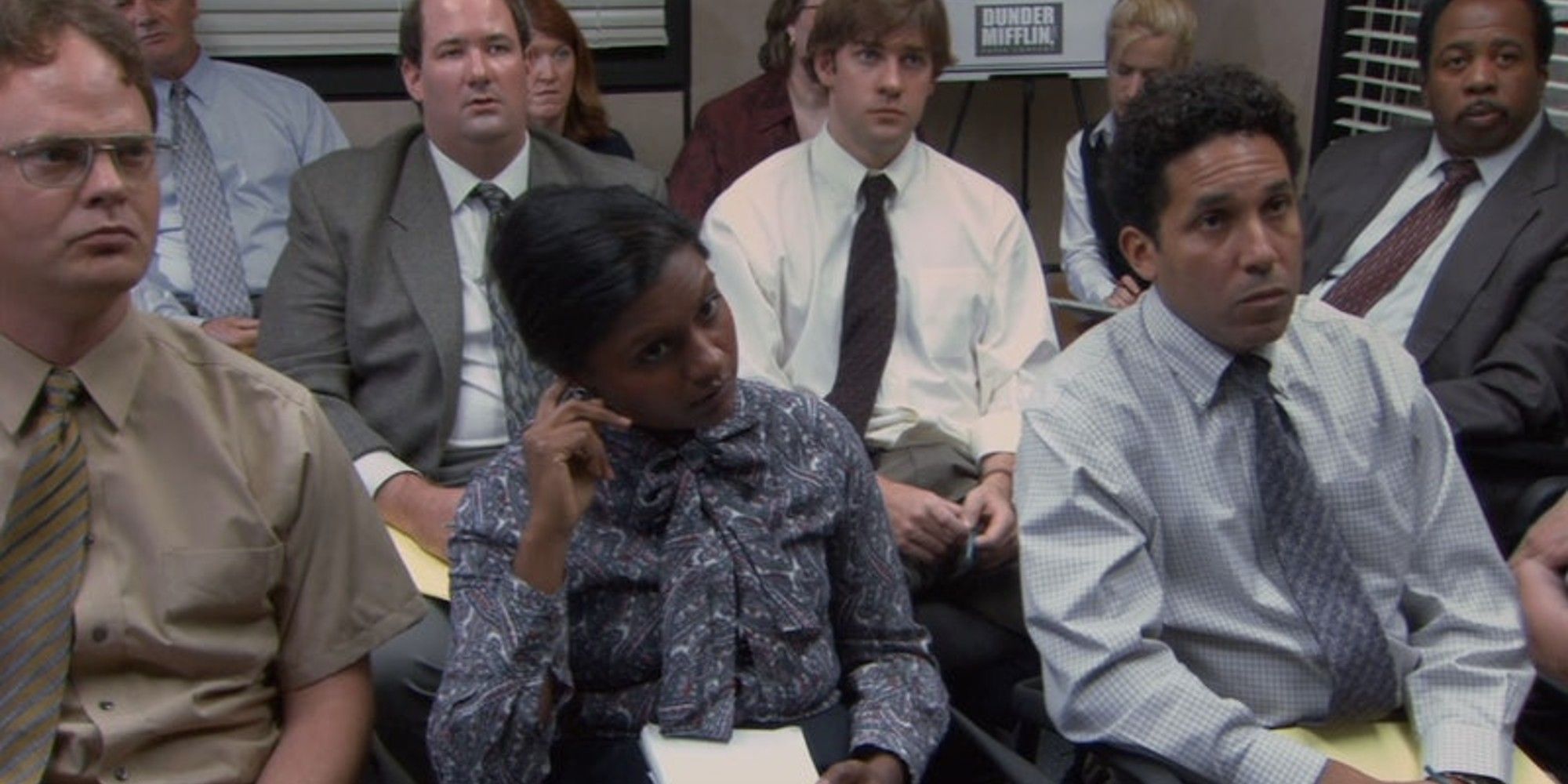 Dwight (Rainn Wilson), Kelly (Mindy Kaling), and Oscar (Oscar Nuñez) sitting in the conference room with the rest of the staff in The Office