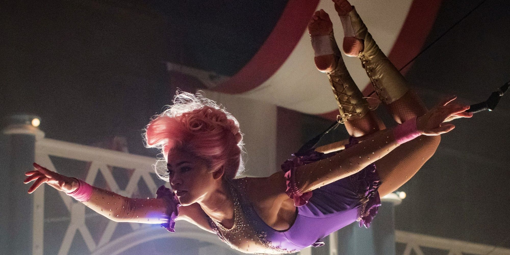 Anne Wheeler dressed in a pink wig and a purple leotard, performs an acrobatic trick under the circus tent in The Greatest Showman.