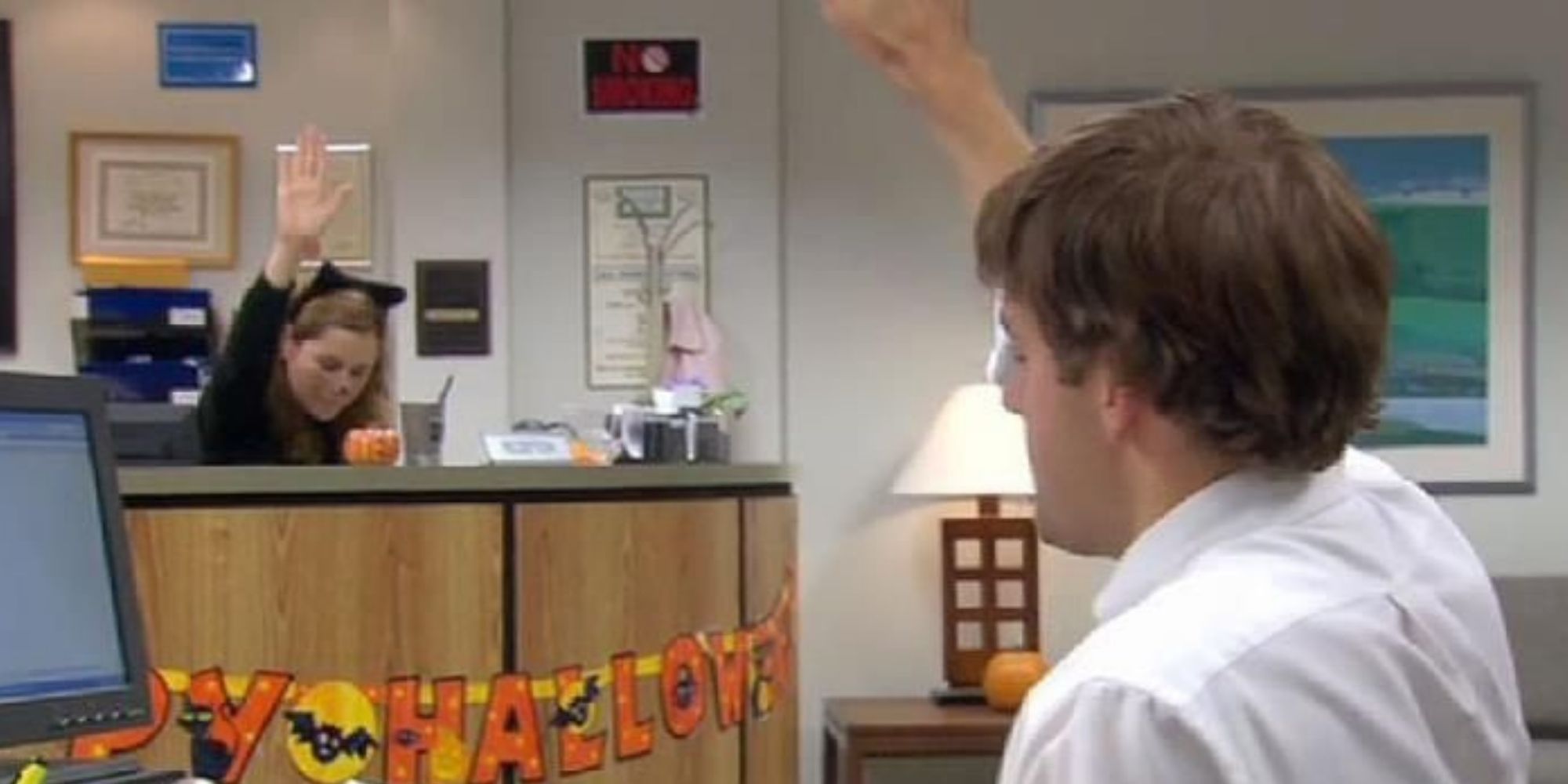 Jim and Pam are high-fiving from far away, and both wearing Halloween costumes. They are not looking at one another, but their computers. Pam is dressed as a cat. The office is decorated for Halloween.