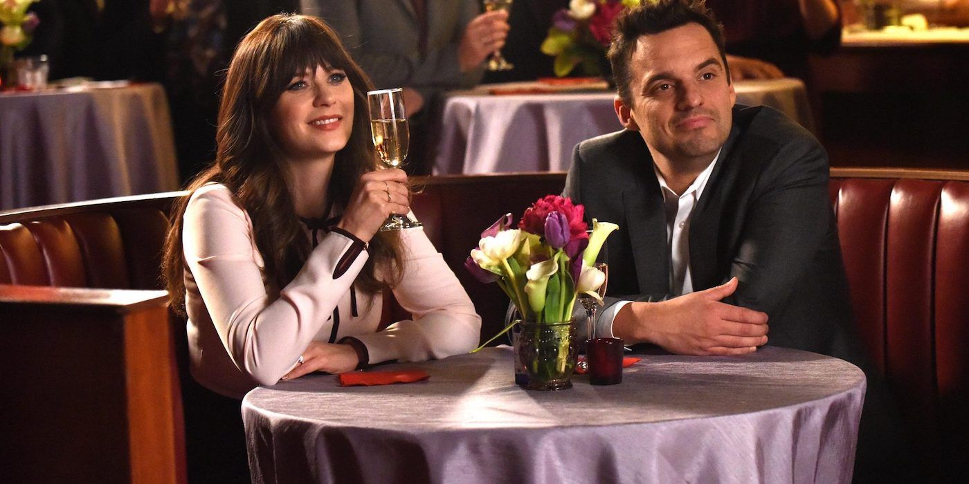 Zooey Deschanel as Jess and Jake Johnson as Nick smiling and sitting at a table in a restaurant in New Girl
