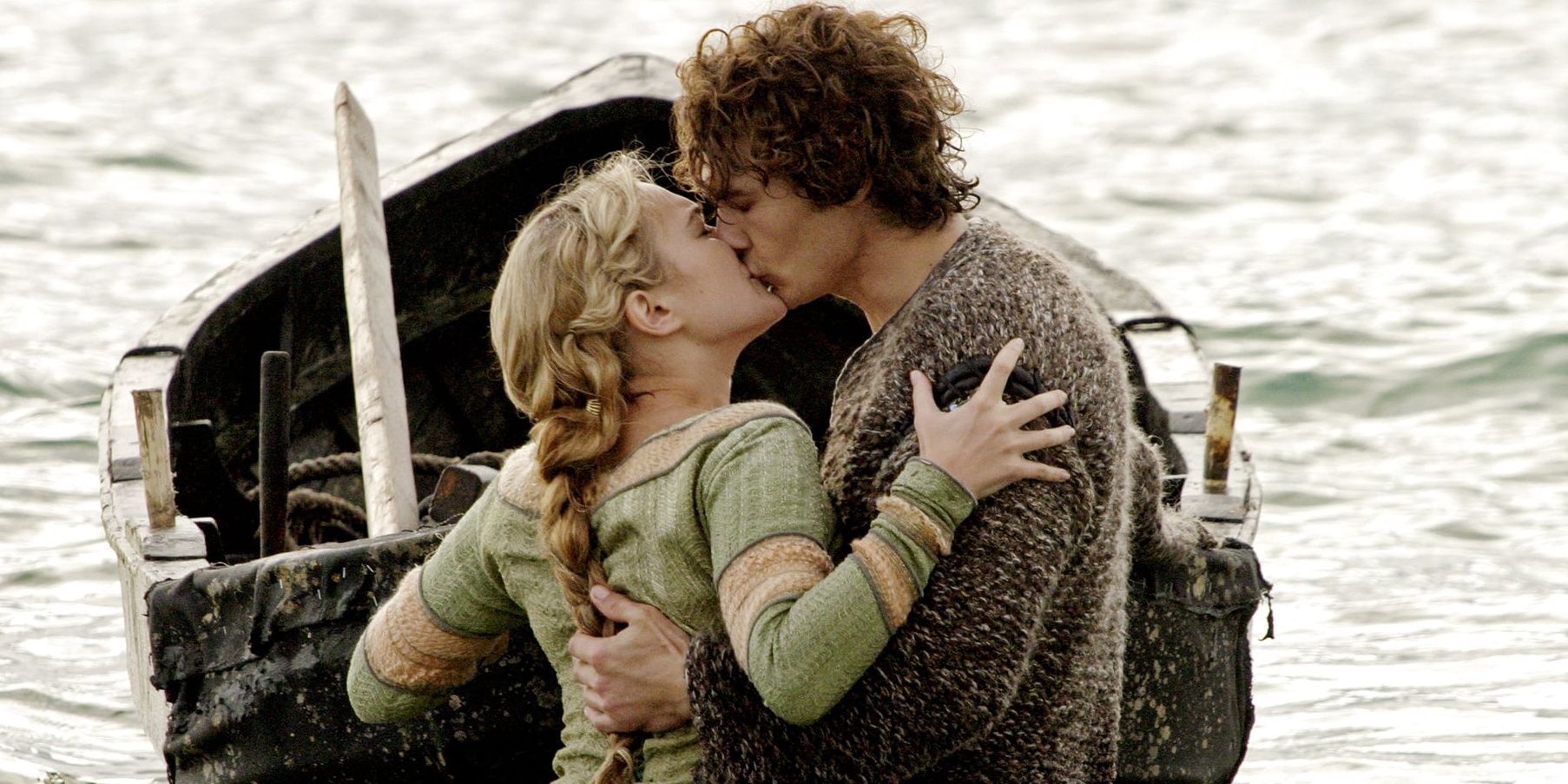 A man and a woman kiss as they stand by a small rowboat by the sea.