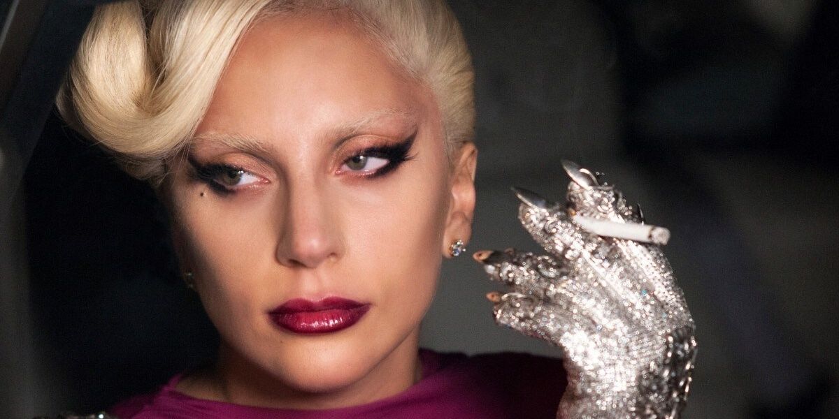 With a bright pink lip and sequin encrusted silver glove, Lady Gaga holds cigarette looking off in American Story Hotel