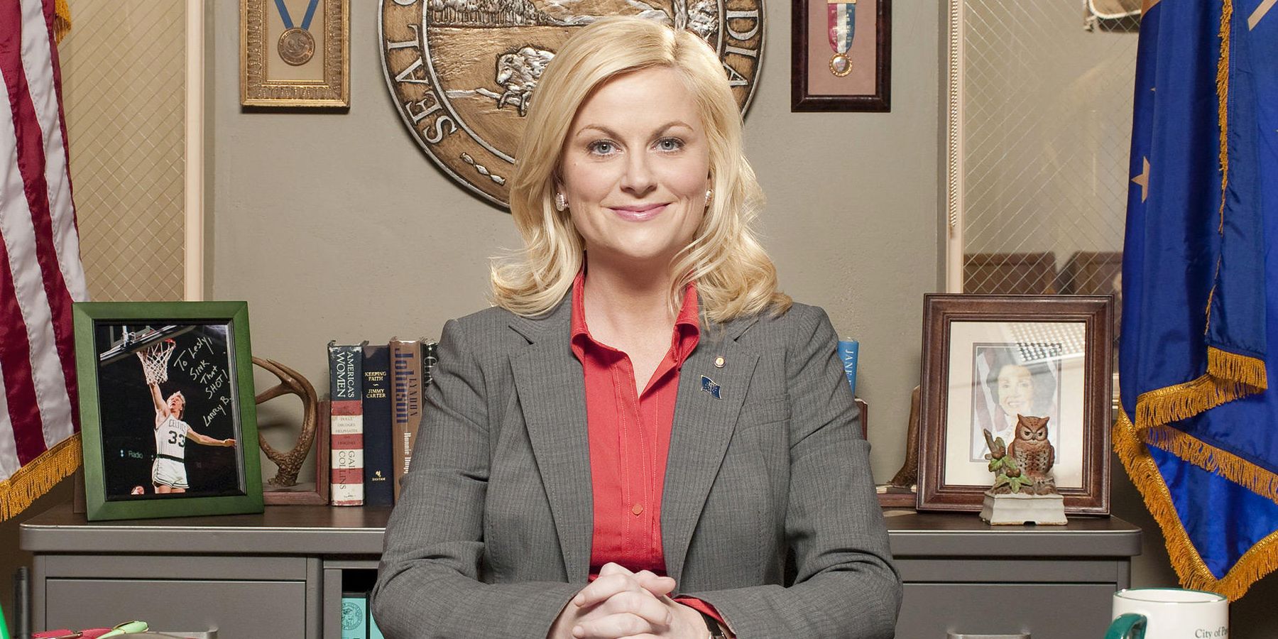 Amy Poehler as Leslie Knope sitting behind her desk and smiling for the camera in Parks and Recreation
