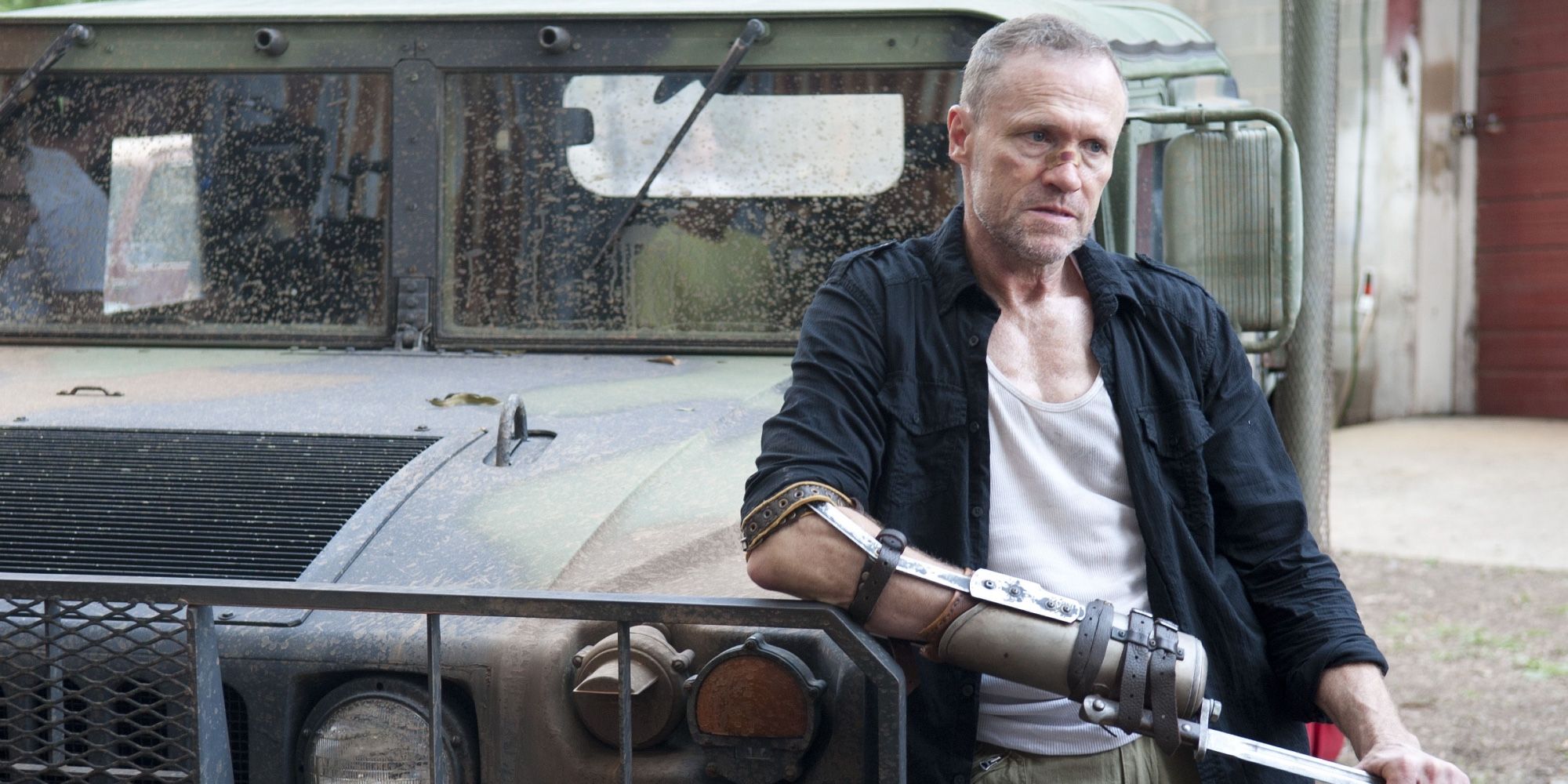 Merle Dixon leans on a military jeep fiddling with his bayonet arm weapon. 