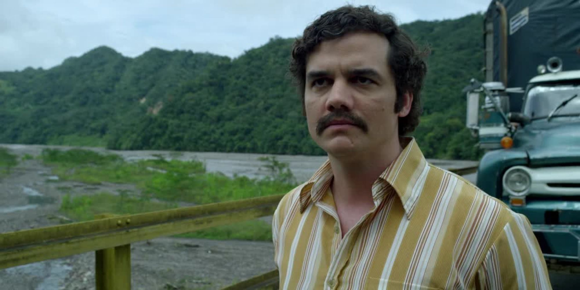 Wagner Moura as Pablo Escobar looks tense in vintage clothing near a vintage truck on a bridge in Narcos.