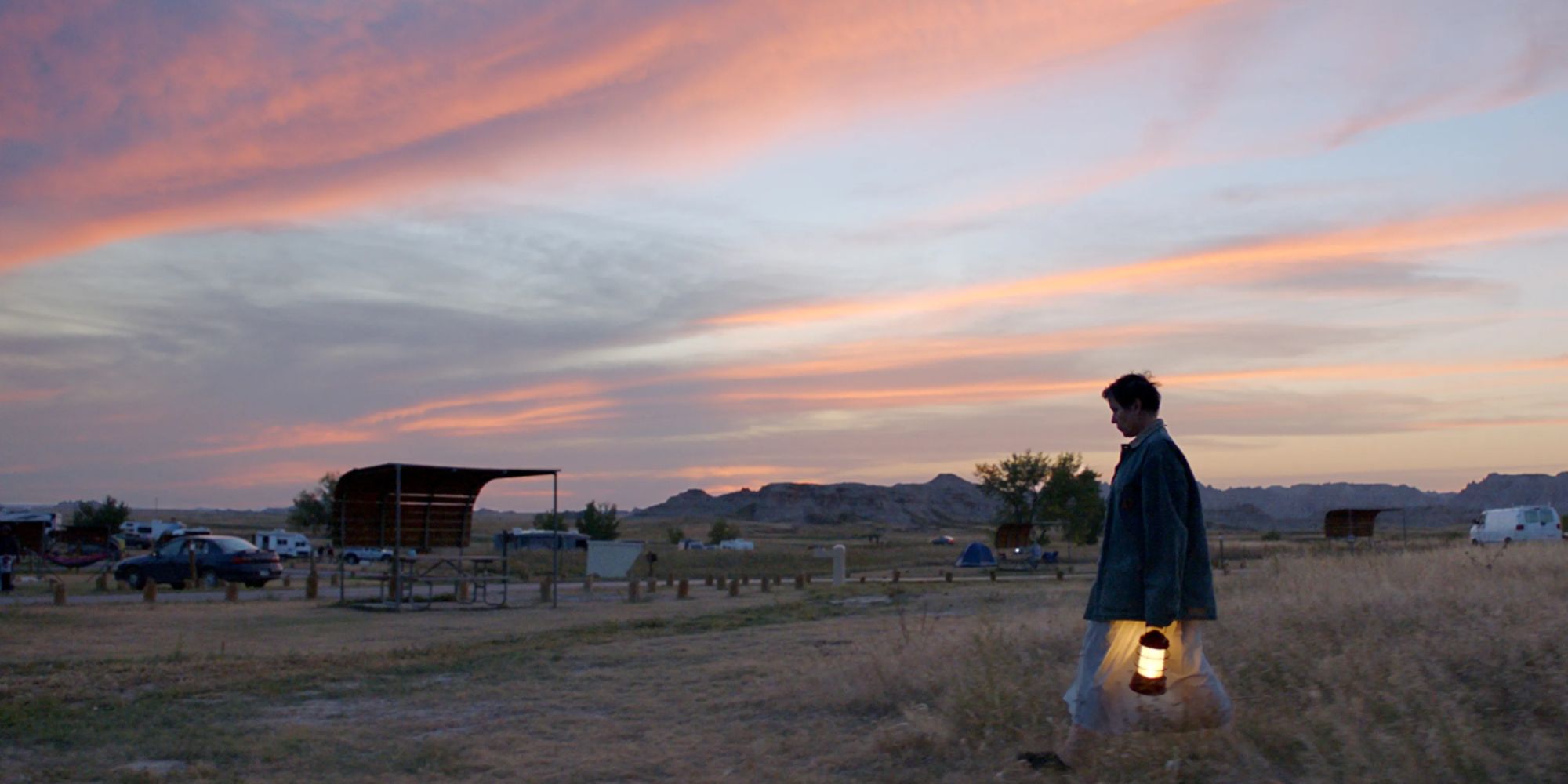 woman walking through a field with a lamp, in front of a sunset