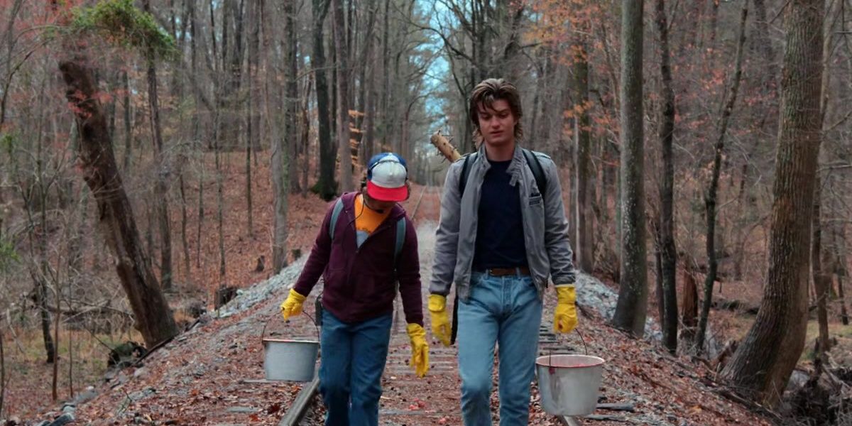 Gaten Matarazzo as Dustin and Joe Keery as Steve walk down the center of train tracks. They carry buckets of meat to lure the monster.