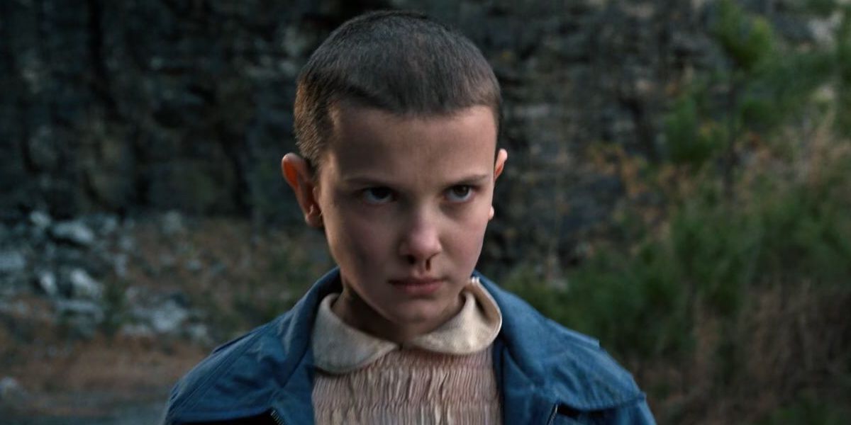 Millie Bobbie Brown as Eleven wears a pink dress with a blue jacket. She looks straight on, using her telekentic powers.
