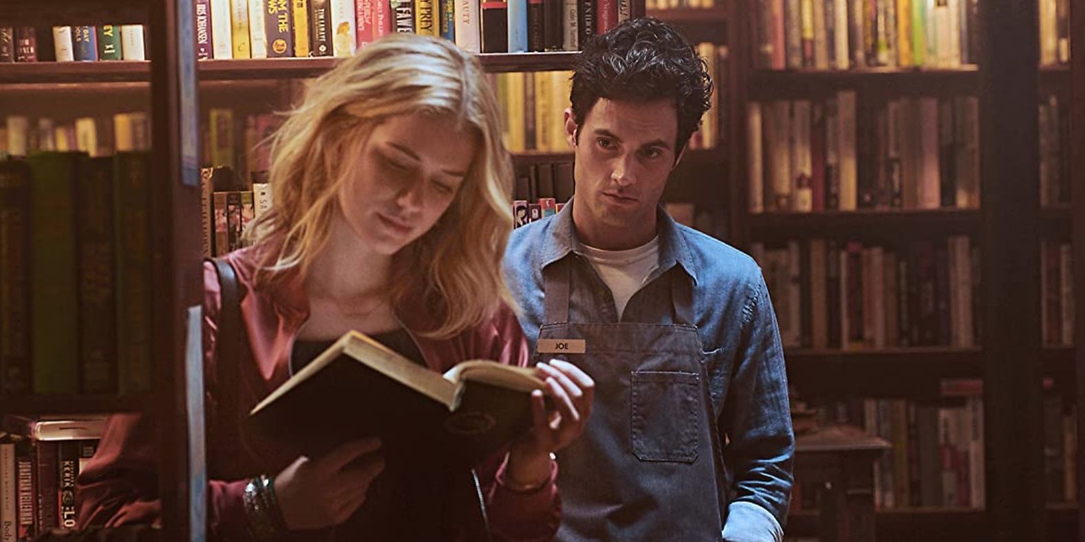 Elizabeth Lail in a library reading a book while Penn Badgley stands behind her in 'You'