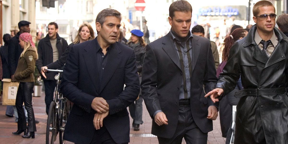 George Clooney como Danny Ocean, Matt Damon como Linus Caldwell e Brad Pitt como Rusty Ryan andando e conversando no meio de uma multidão em Oceans Twelve