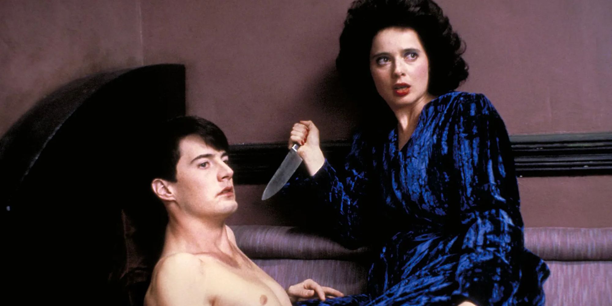 A woman in a blue velvet robe holds a knife pointed at a shirtless man 