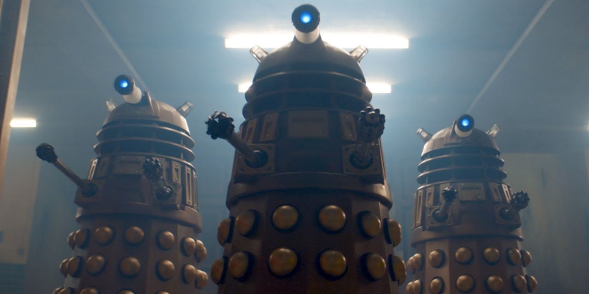 Three Daleks stand over the camera in a dimly lit corridor