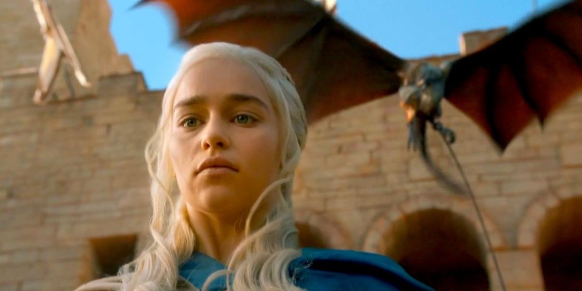 Emilia Clarke as Daenerys Targaryen in a blue robe stands before a temple with a chained screeching dragon flying behind her.