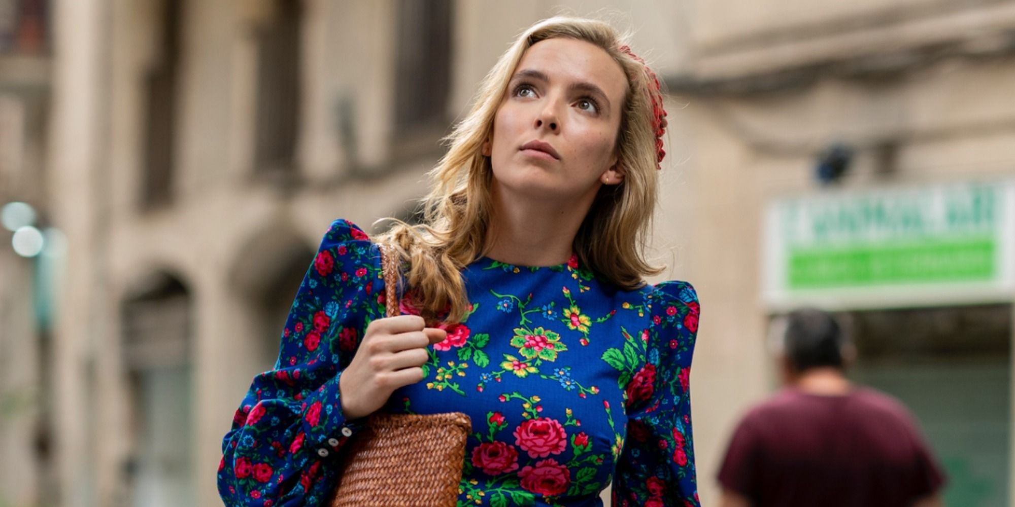 Jodie Comer as Villanelle in Killing Eve walking around holding a purse in a floral dress, looking up.