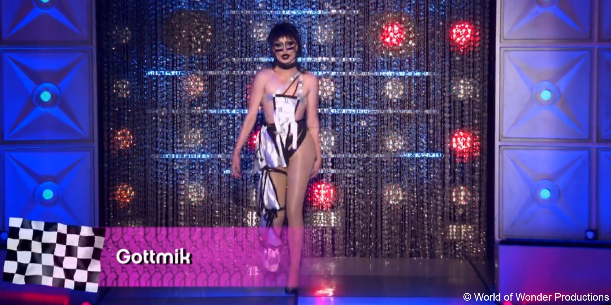 A still of drag queen Gottmik wearing a strappy black and silver garment with grey sunglasses