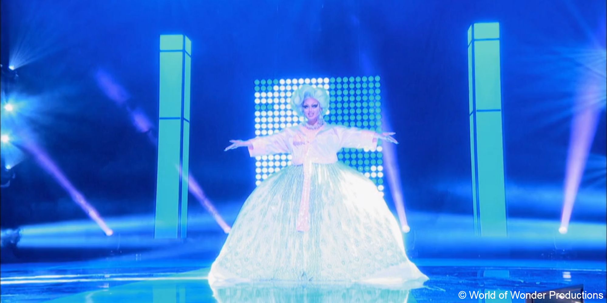 A still of drag queen Kim Chi wearing a luminescent blue and white ball gown, arms spread wide