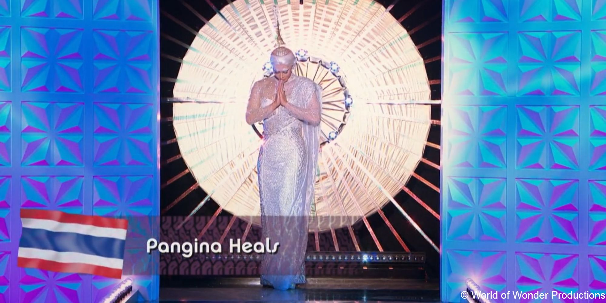 A still of drag queen Pangina Heals wearing a beaded white gown and Thai-inspired headdress, bowing