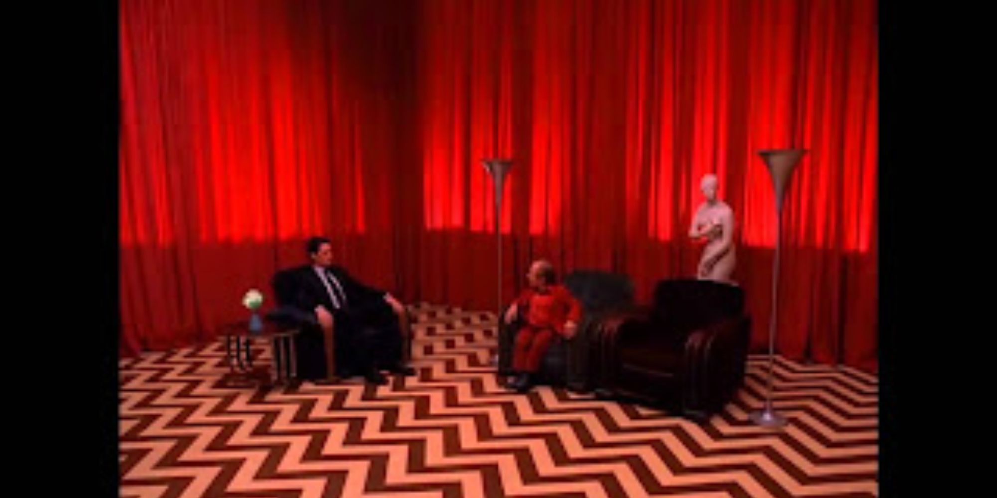 A man in a suit and a little person sit on couches in a room lined by a red curtain with a chevron patterned floor