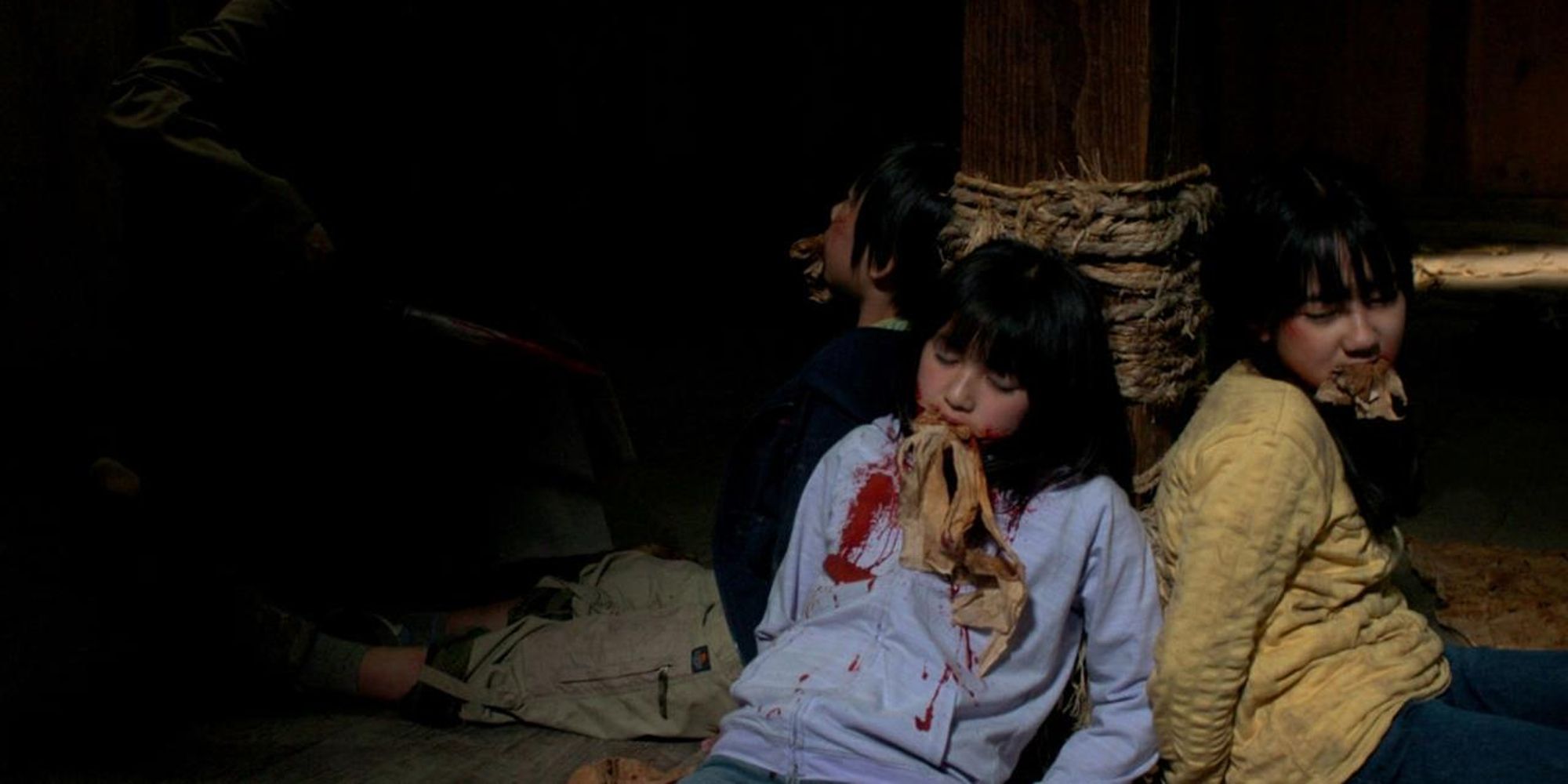 Two women and a man sit tied to a post in a basement with bloodied shirts and rags shoved into their mouths.