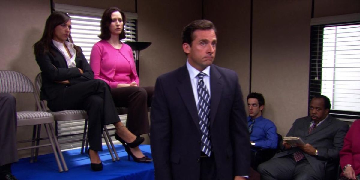 Michael Scott from The Office stands in front of two employees, who are seated on chairs on top of a table