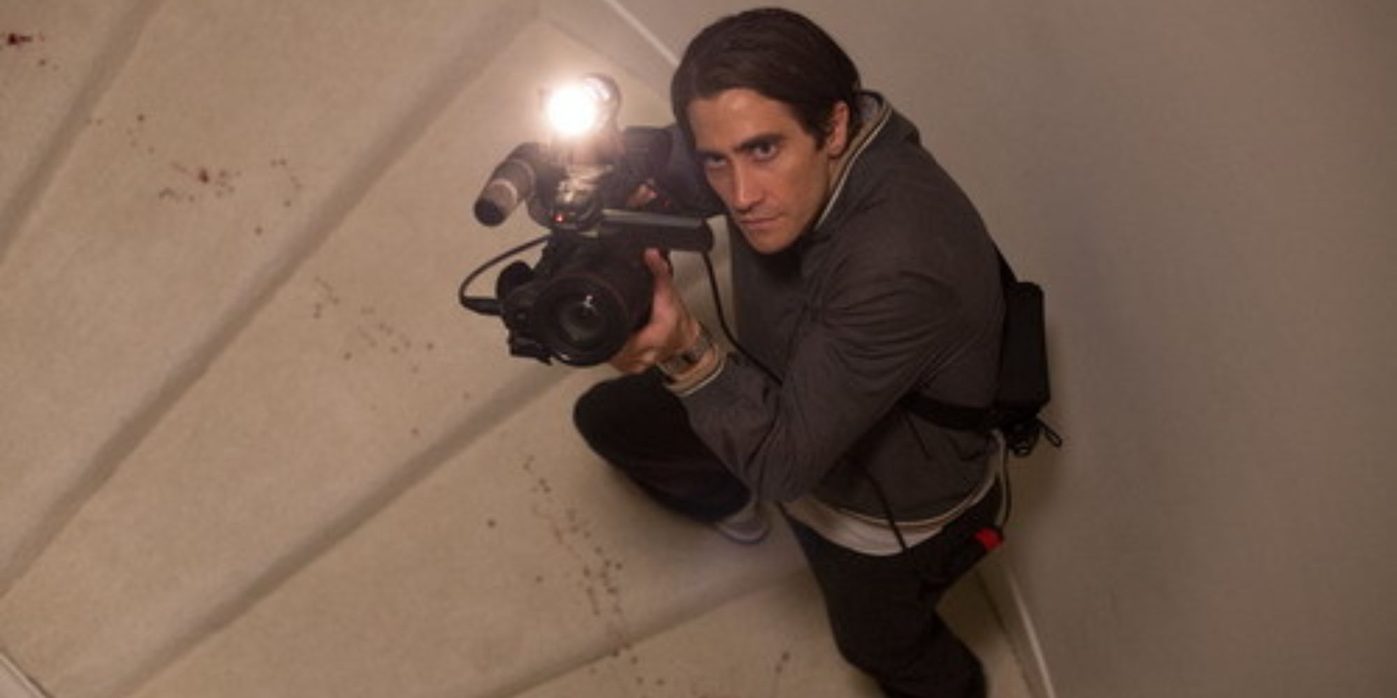 Jake Gyllenhaal dans le rôle de Lou Bloom montant les escaliers en tenant une caméra dans 'Nightcrawler'.