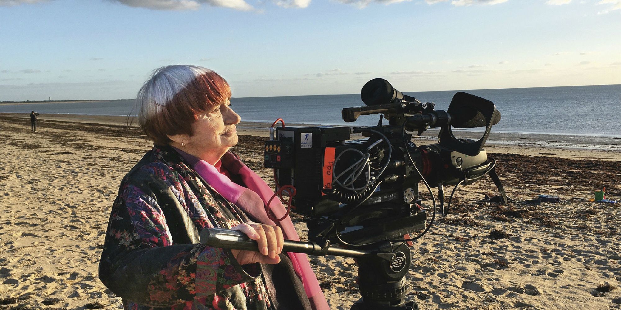 Agnes Varda di pantai dengan kamera memandang ke kejauhan.