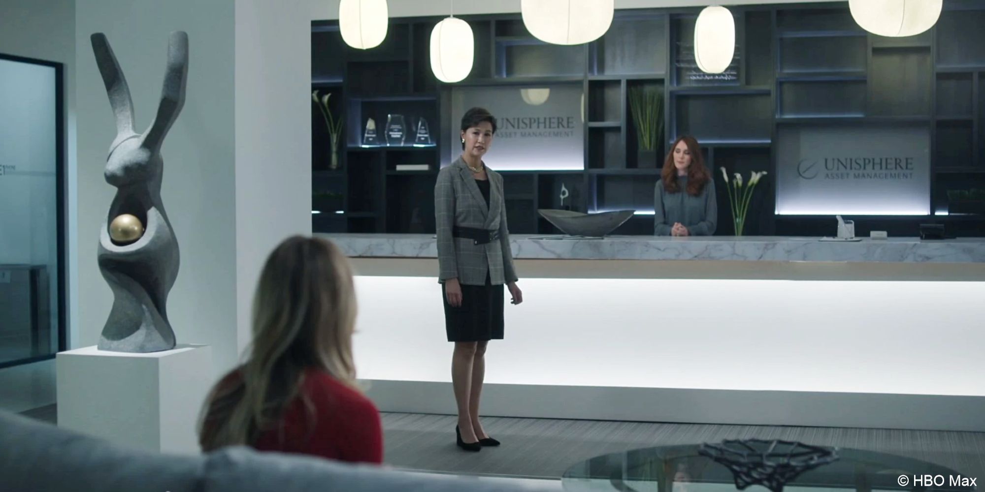 Cassie sits on a couch at Unisphere, back to the camera, as two businesswomen look at her.