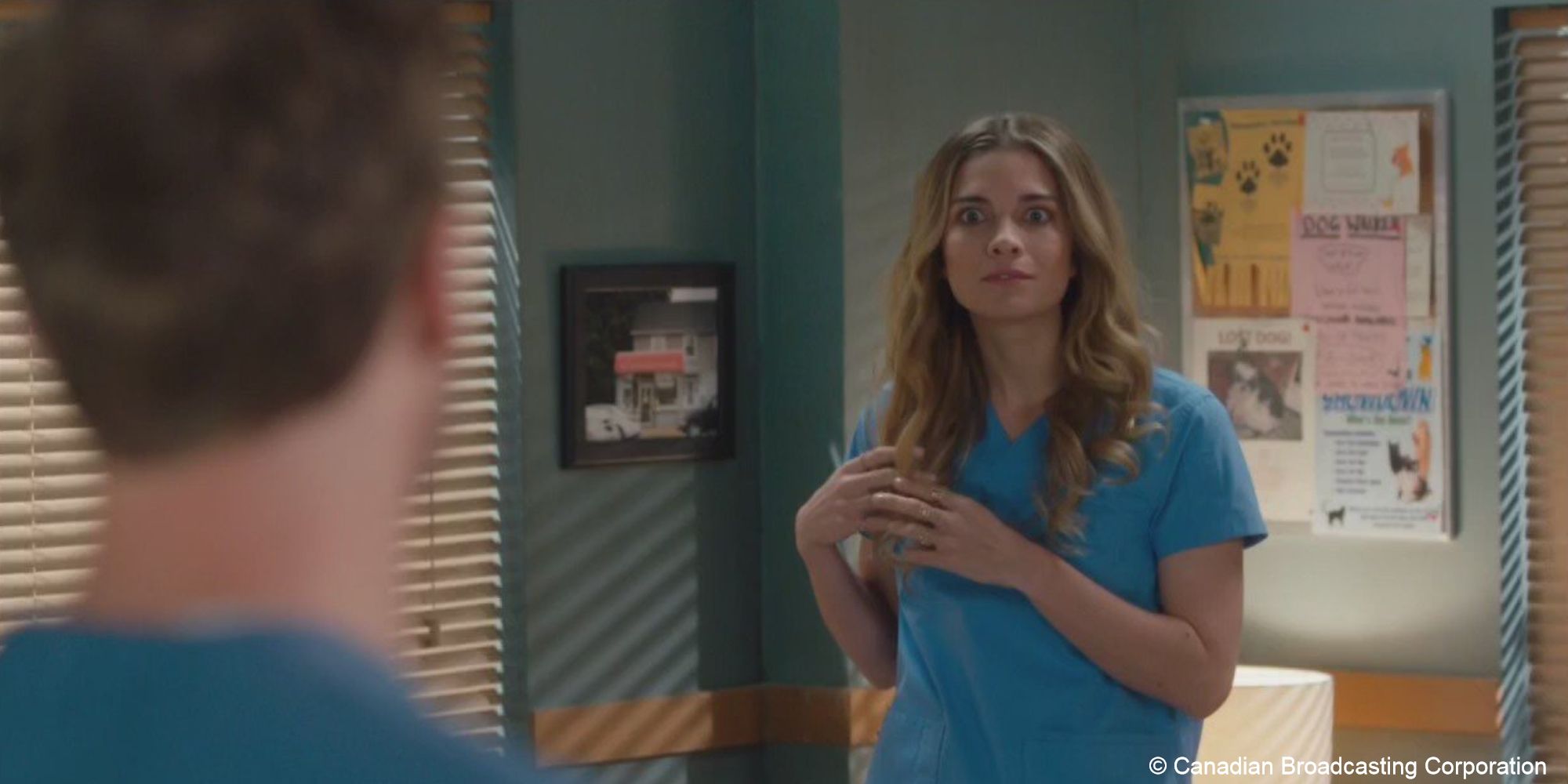 Alexis stands in scrubs, twirling her hair as she looks at Ted whose back is to the camera