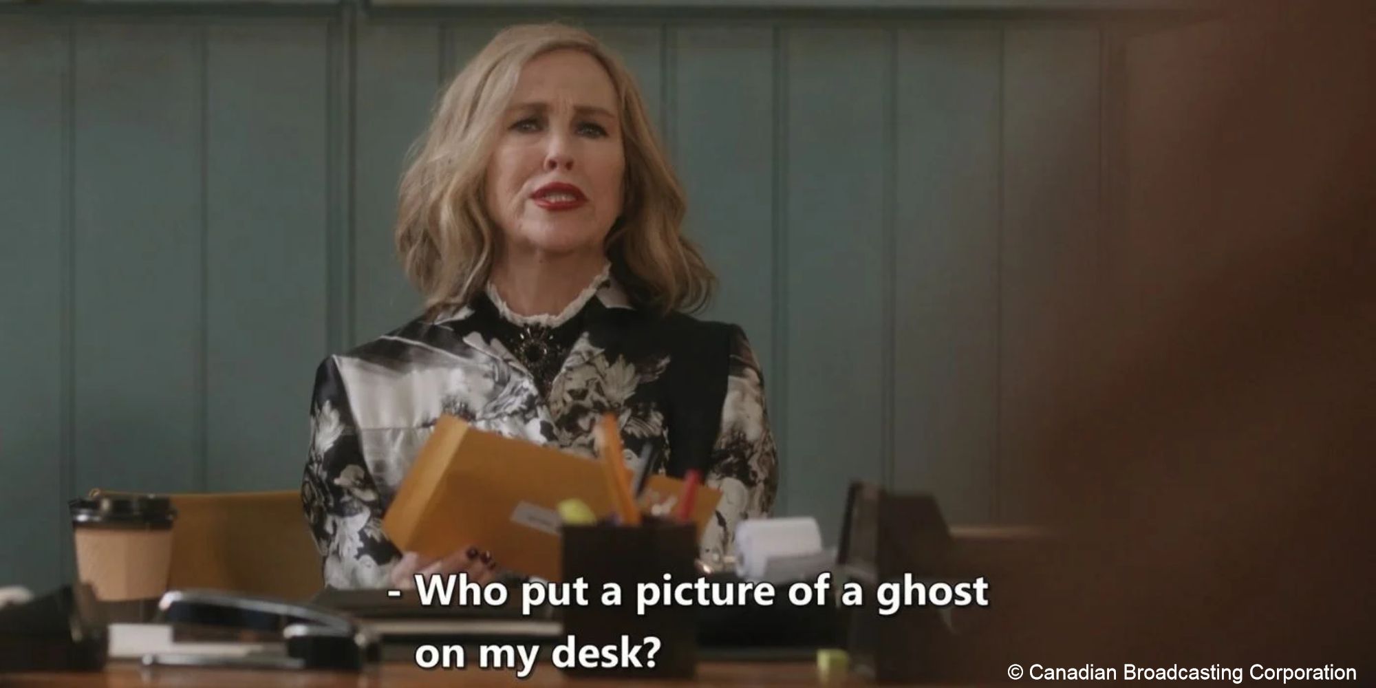 Moira opens a manila envelope at her desk, caption reads "who put a picture of a ghost on my desk?"
