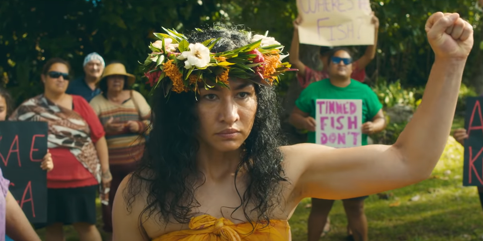 The character Vai from 'Vai' raises her left fist in defiance. She wears a flowered lei on her head.