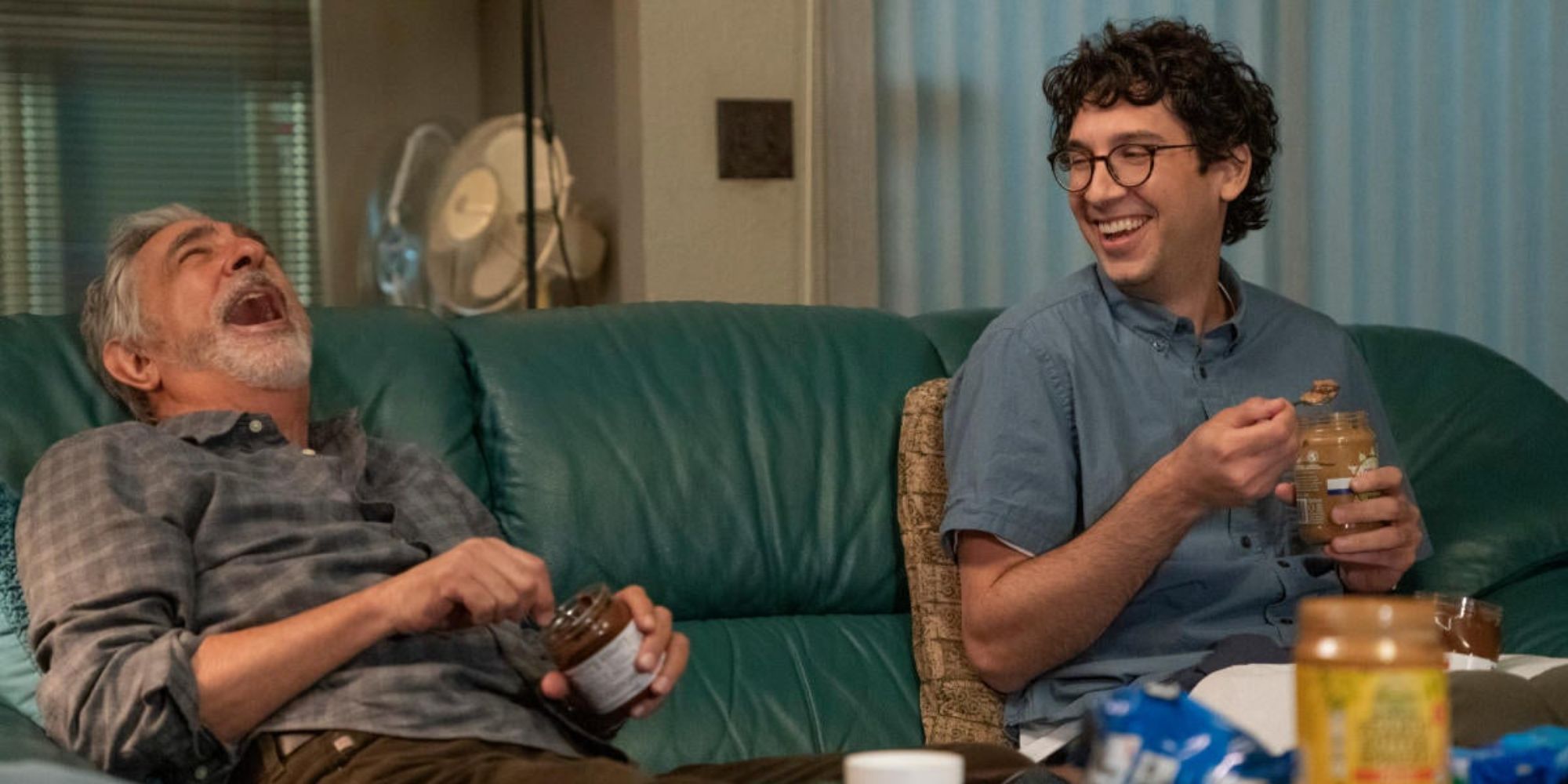 An old man and a young one sitting on the couch eating peanut butter from the jars and laughing