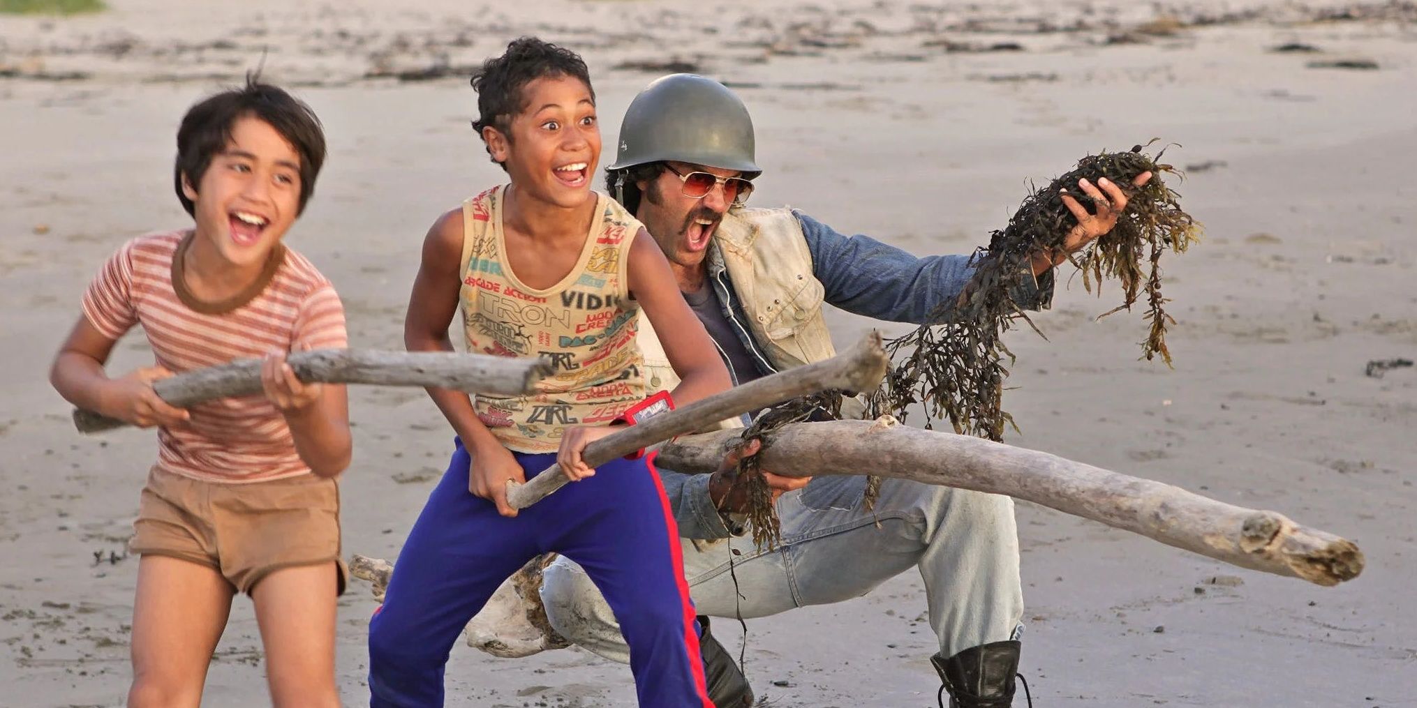 The characters Rocky, Boy, and Alamein from the film 'Boy' stand on a beach with driftwood.