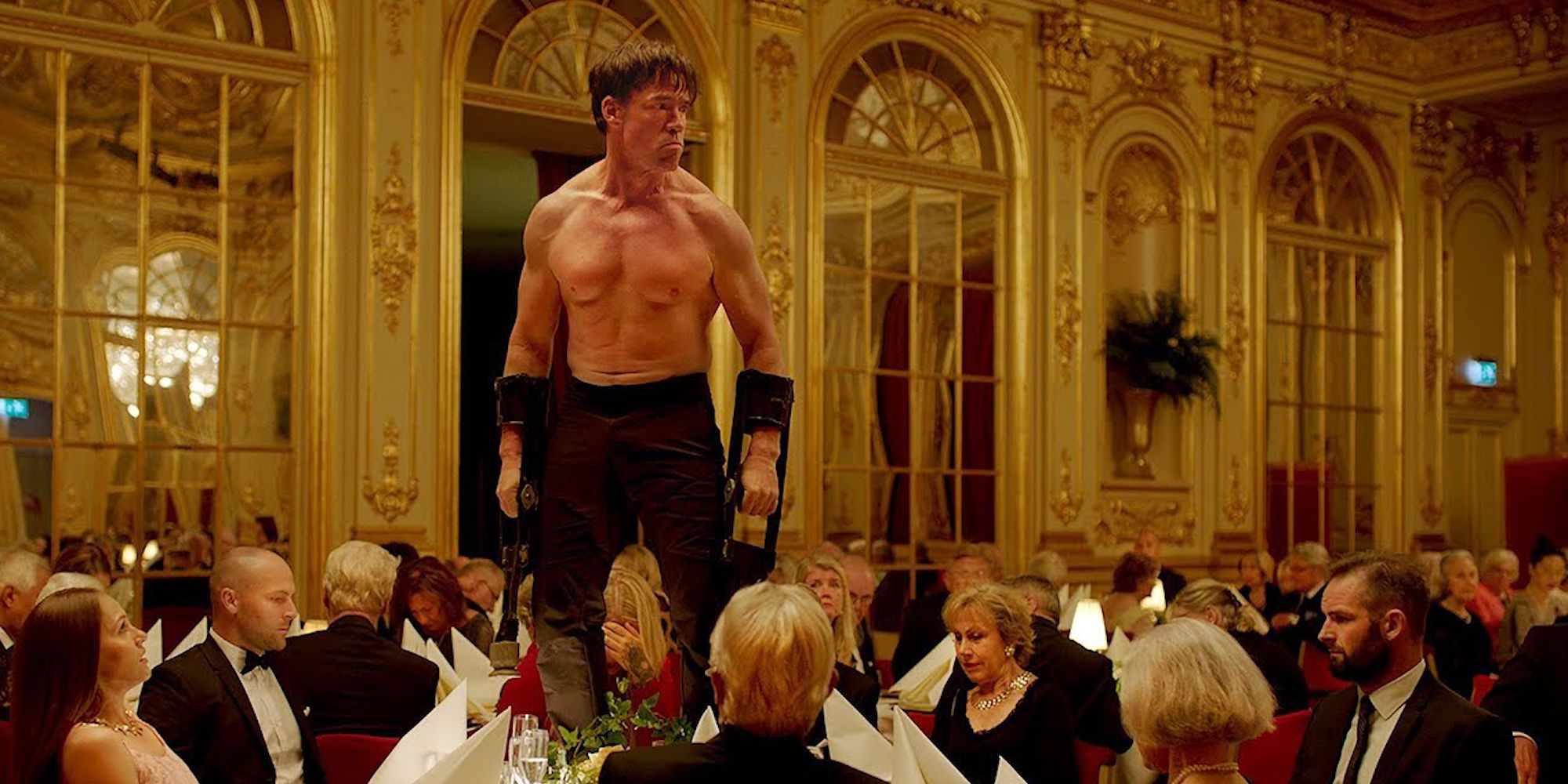 A shirtless man with arm contraptions stands at a black tie function. 