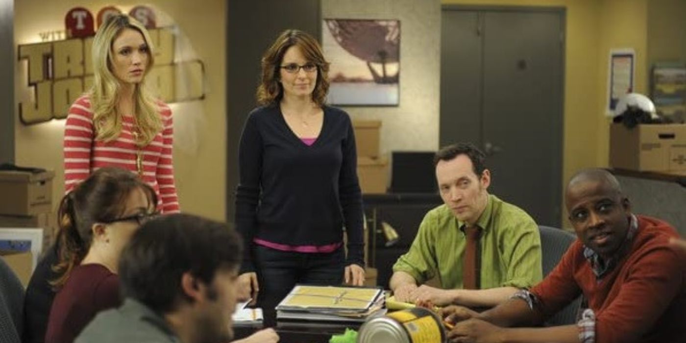 Tina Fey and others from the team sitting around a desk in a writer's meeting in a scene from 30 Rock.