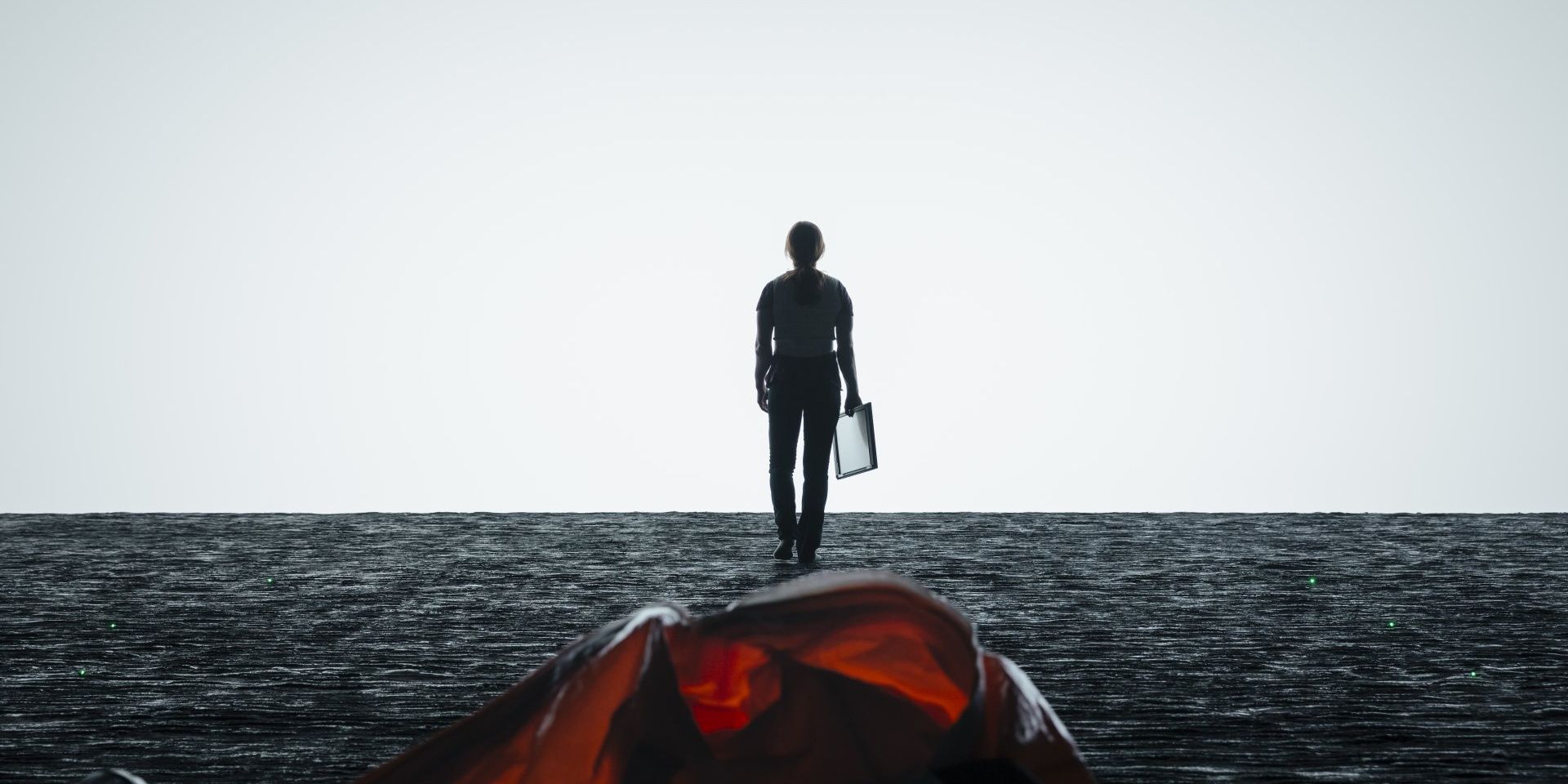 Amy Adams as Louise Banks in Arrival walking across a gray, rocky floor toward a blank white expanse. In the foreground, her orange hazmat suit lies discarded on the floor and she carries a clipboard as she walks.