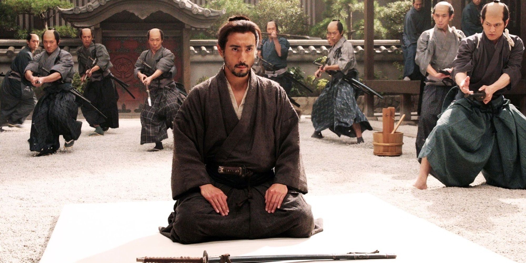 A samurai sits while others raise their swords at him in Hara-Kiri Death of a Samurai