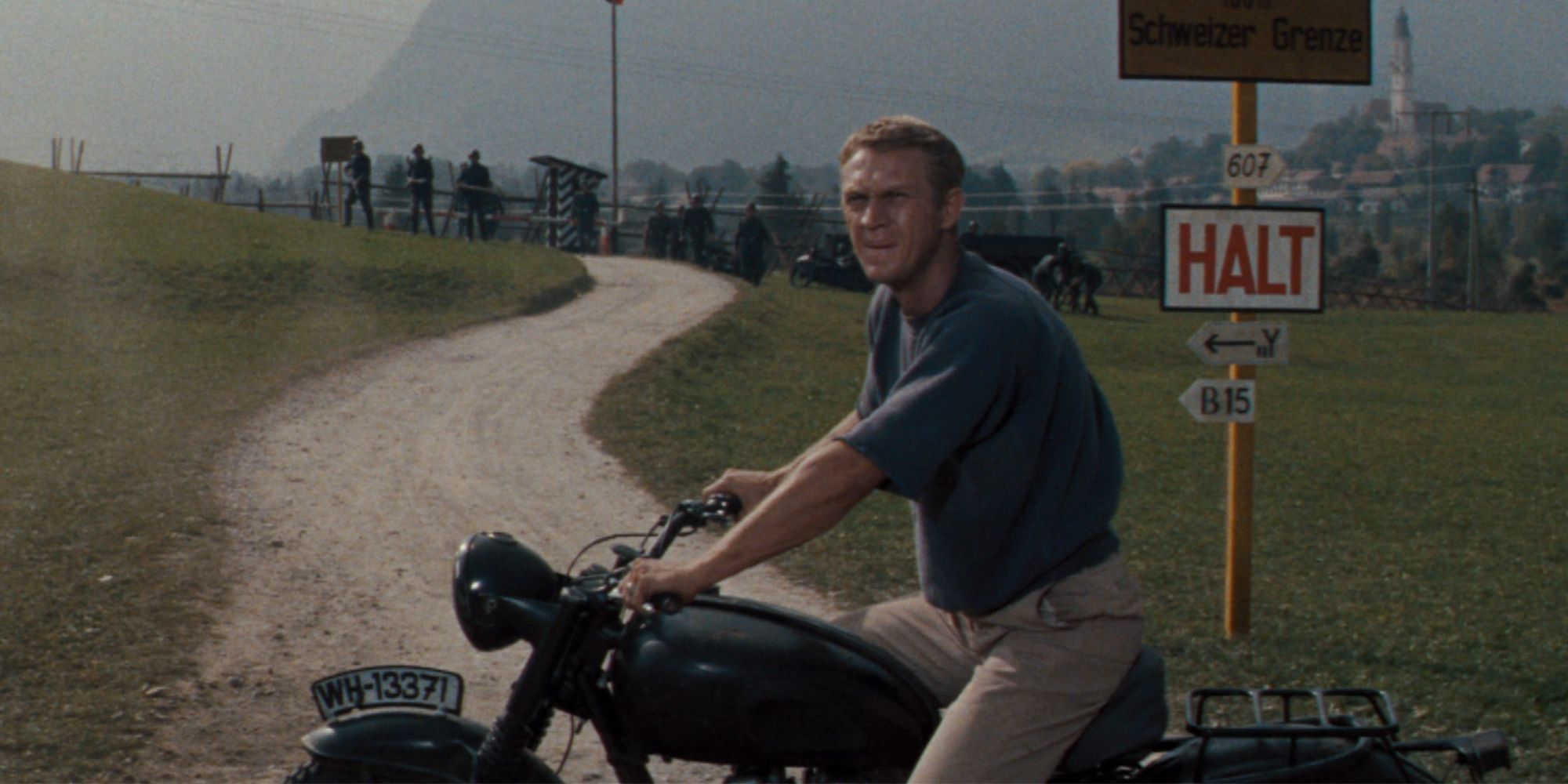 Steve McQueen riding a motorcycle stopped along a dirt road outside a prison camp in The Great Escape