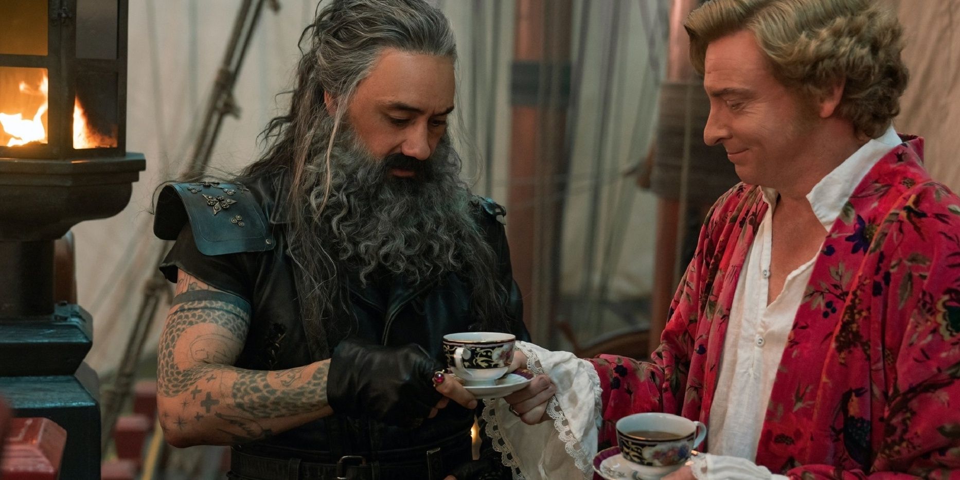 Stede Bonnet, wearing a floral pink robe, hands Blackbeard, wearing a black motorcycle jacket, a teacup.