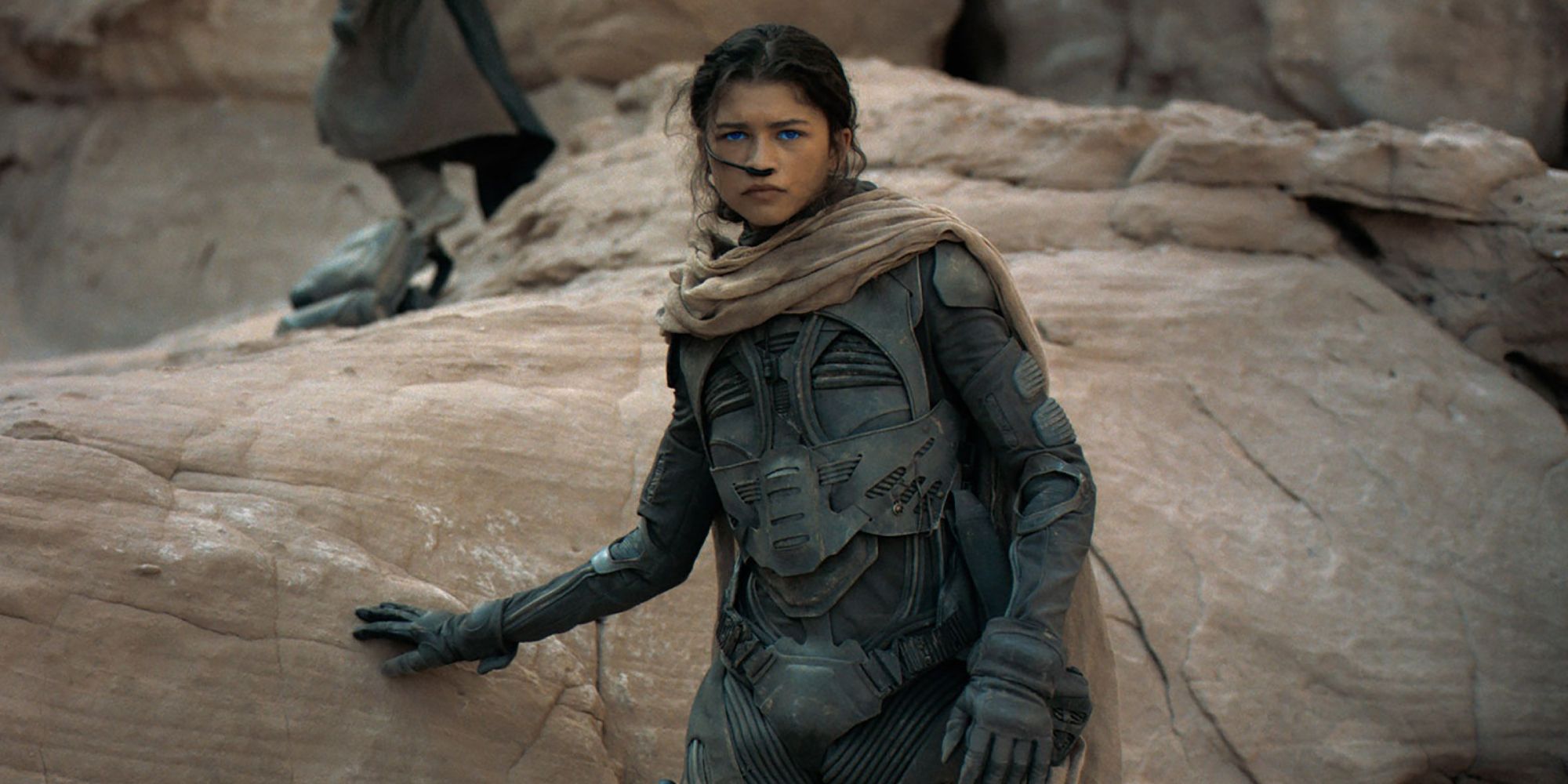 Chani, wearing a stillsuit, leaning against a rock in Dune.