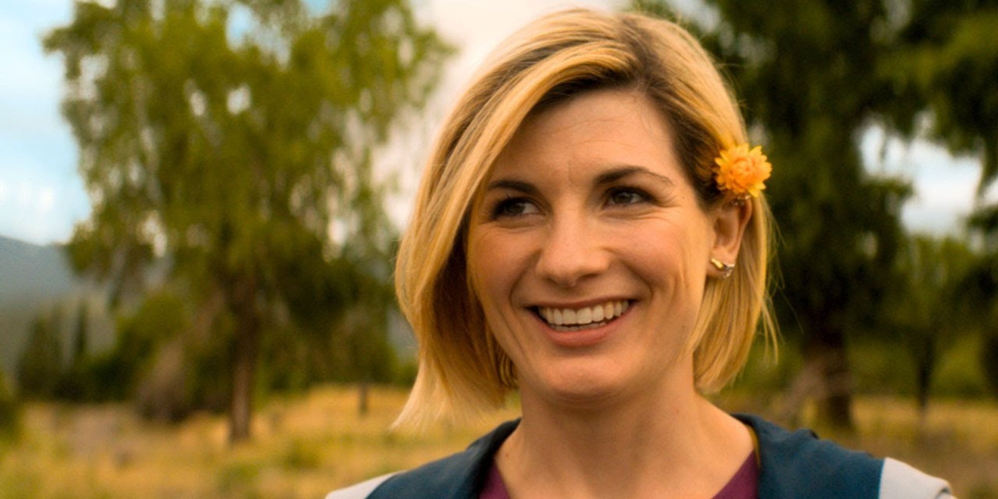 Jodie Whittaker playing the Thirteenth Doctor with a flower in her hair smiling in grasslands