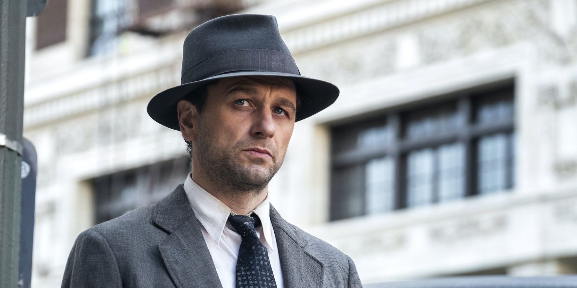 Matthew Rhys as Perry Mason in a suits and hat looks troubled standing outside in Perry Mason.