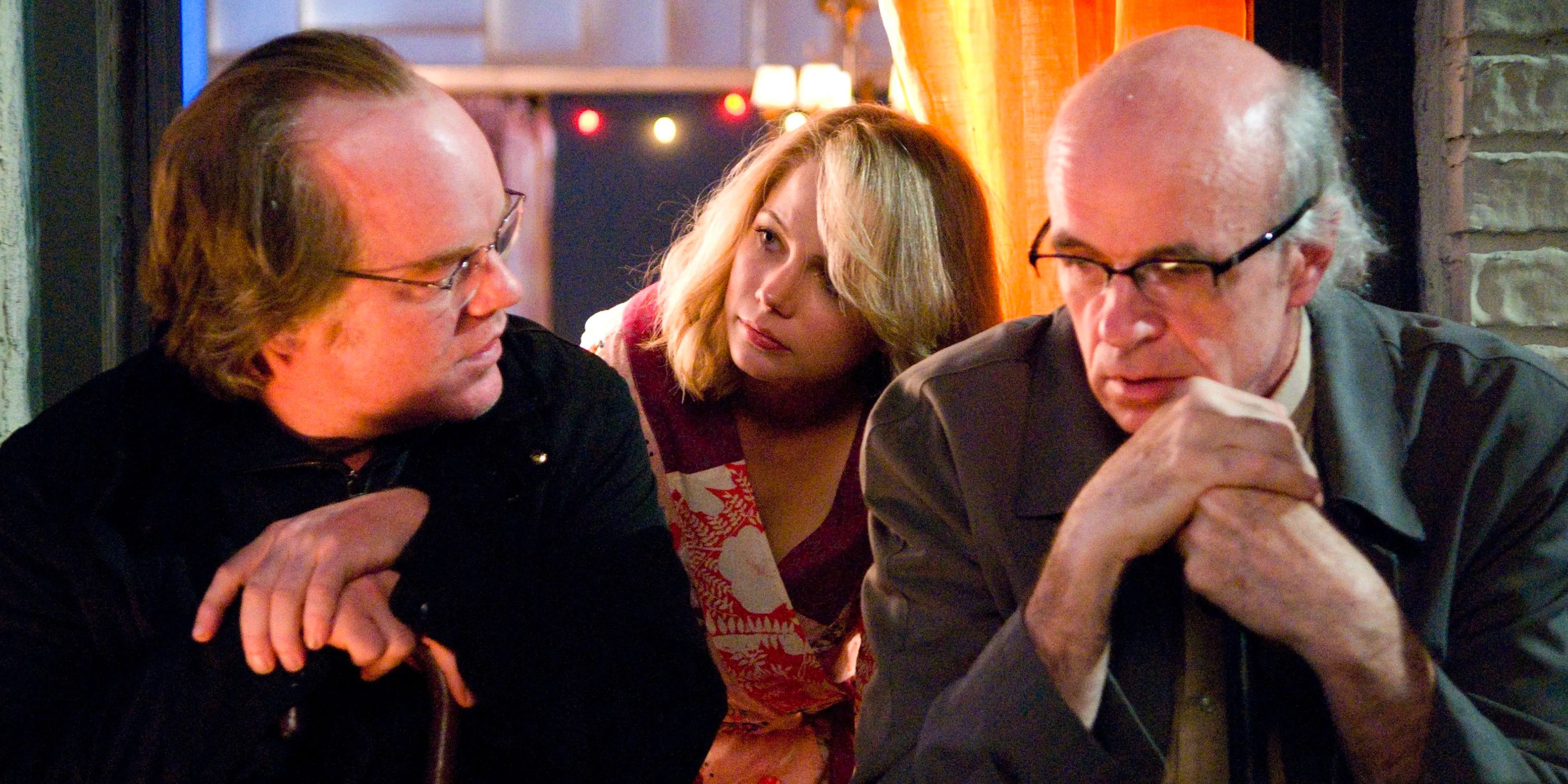 Three people sit outside a window of a New York apartment and talk. 