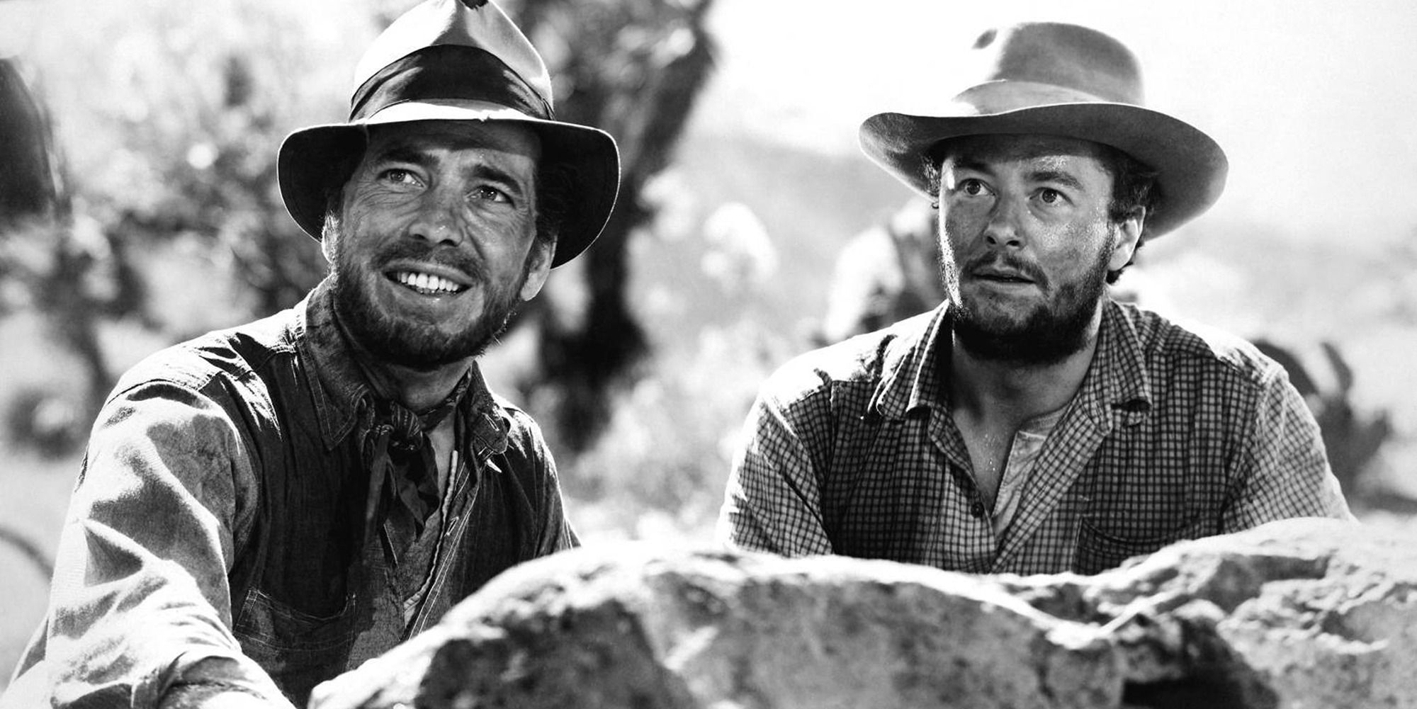 Two men smiling while sitting behind a rock, wearing traditional Western movie clothes